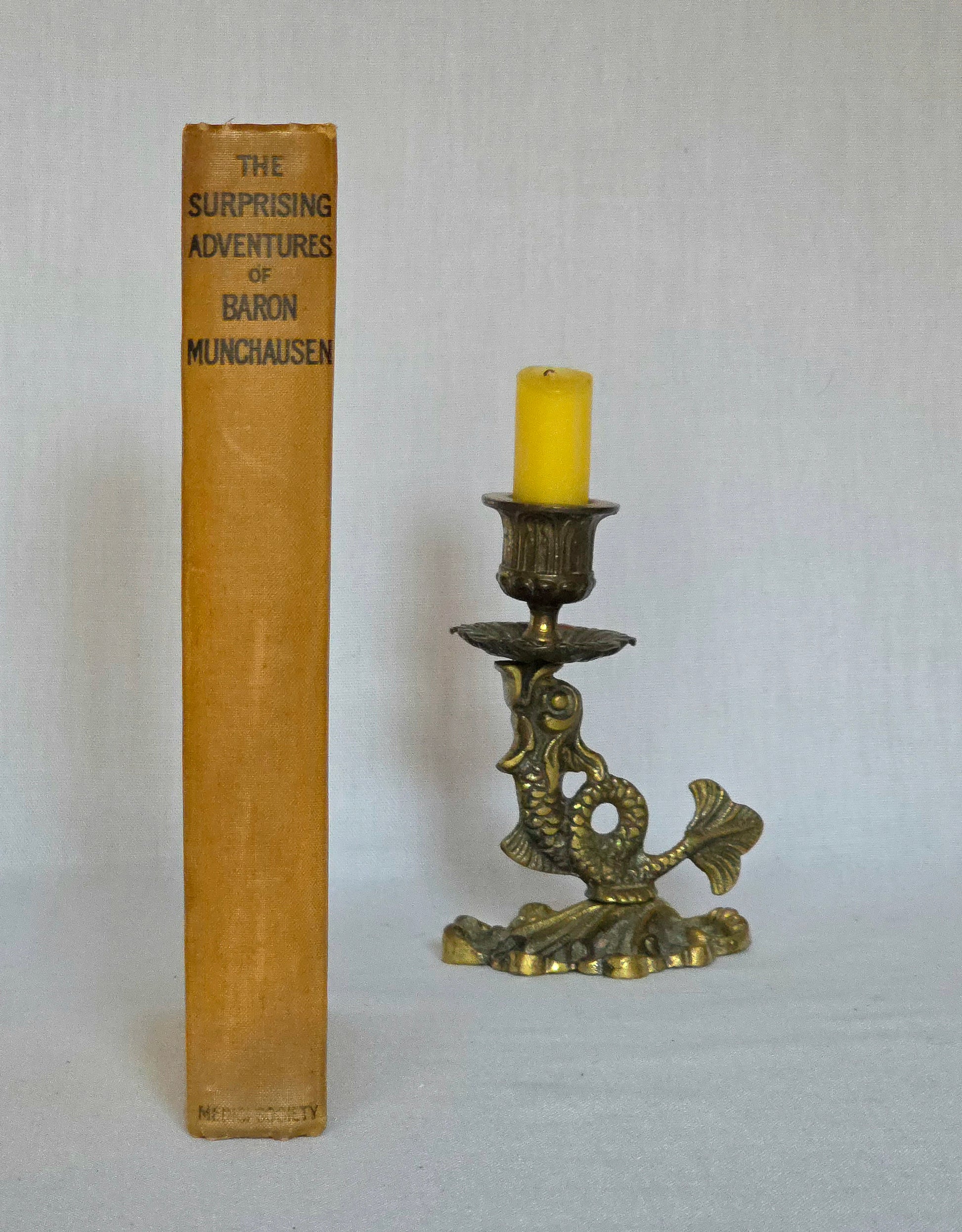 Yellow spine of vintage book titled 'The Surprising Adventures of Baron Munchausen' next to a decorative brass candlestick with a candle.
