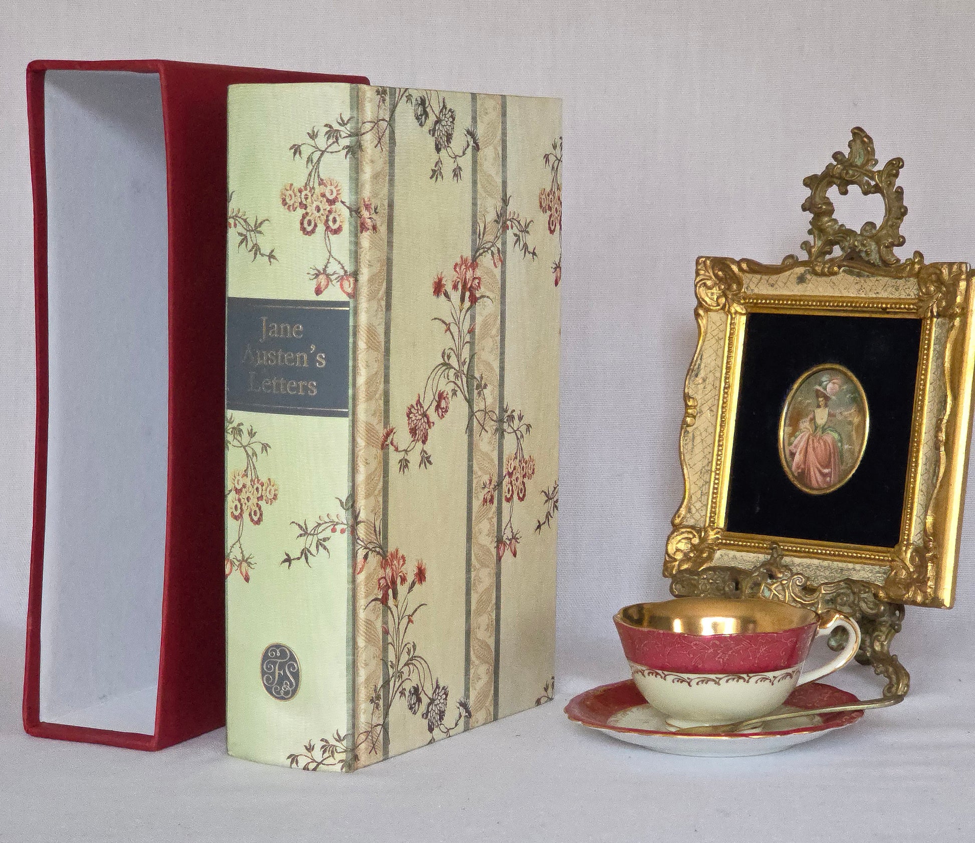 Decorative book being a copy of Jane Austen's Letters, The Folio Society a teacup and saucer, and an ornate gold frame on a white background.