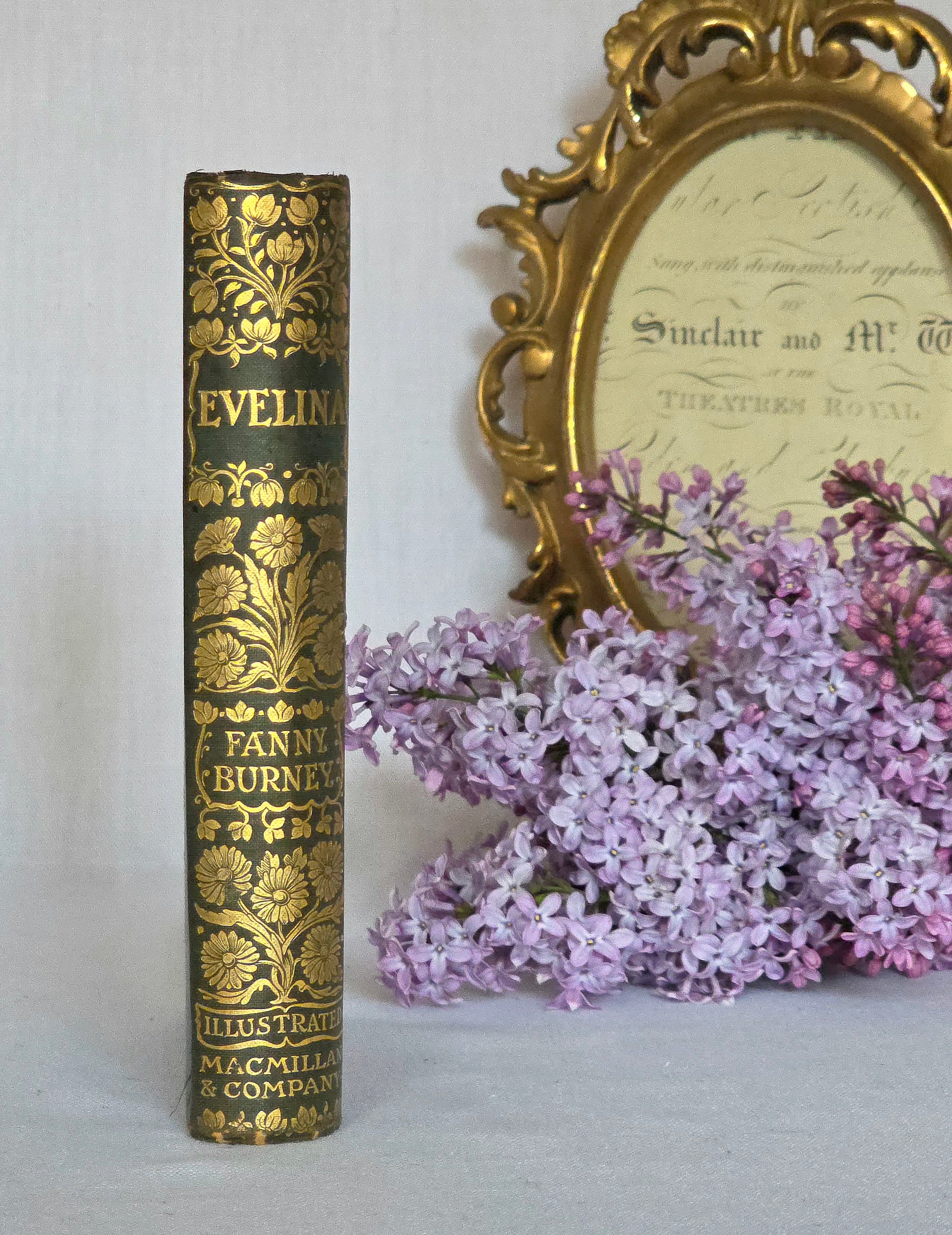 Vintage book spine titled 'Evelina' by Fanny Burney with a gold decorative cover, next to a bouquet of lilacs and an ornate gold frame.