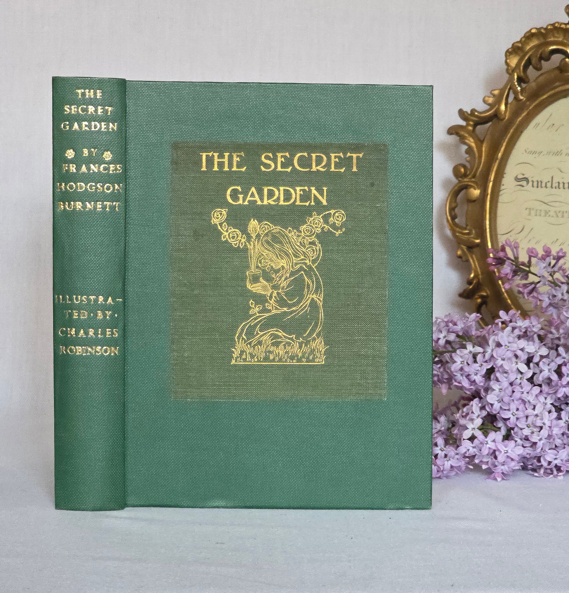 Antique book titled 'The Secret Garden' by Frances Hodgson Burnett with decorative cover and gold text, placed on a white surface with a decorative frame and flowers in the background.