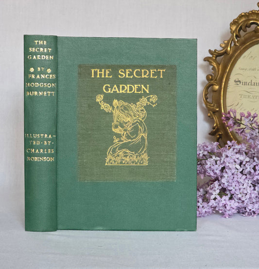 Antique book titled 'The Secret Garden' by Frances Hodgson Burnett with decorative cover and gold text, placed on a white surface with a decorative frame and flowers in the background.