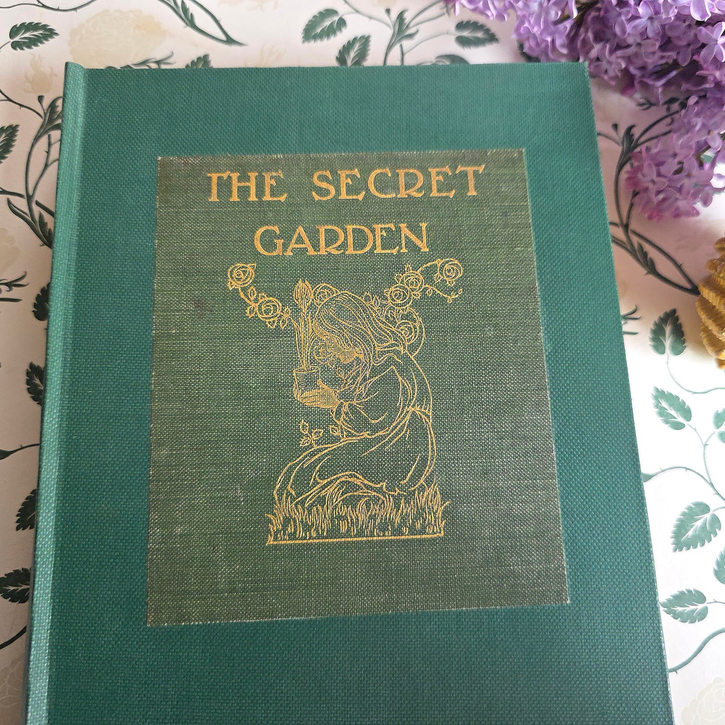 Antique book titled 'The Secret Garden' by Frances Hodgson Burnett with decorative cover and gold text, placed on a white surface with a decorative frame and flowers in the background.