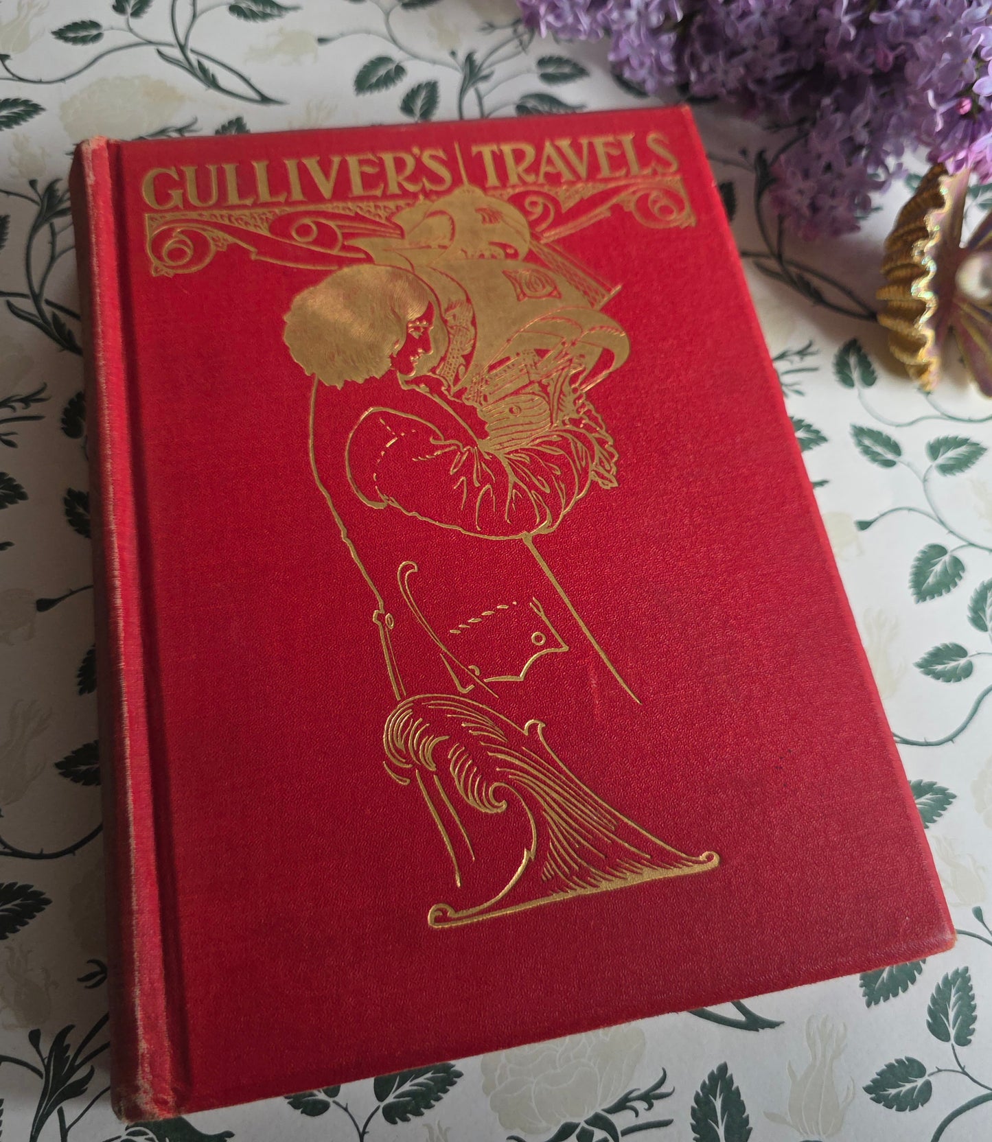 Red book titled 'Gulliver's Travels' with gold illustrations on a light gray background. Illustrated by AE Jackson and published by Nister.