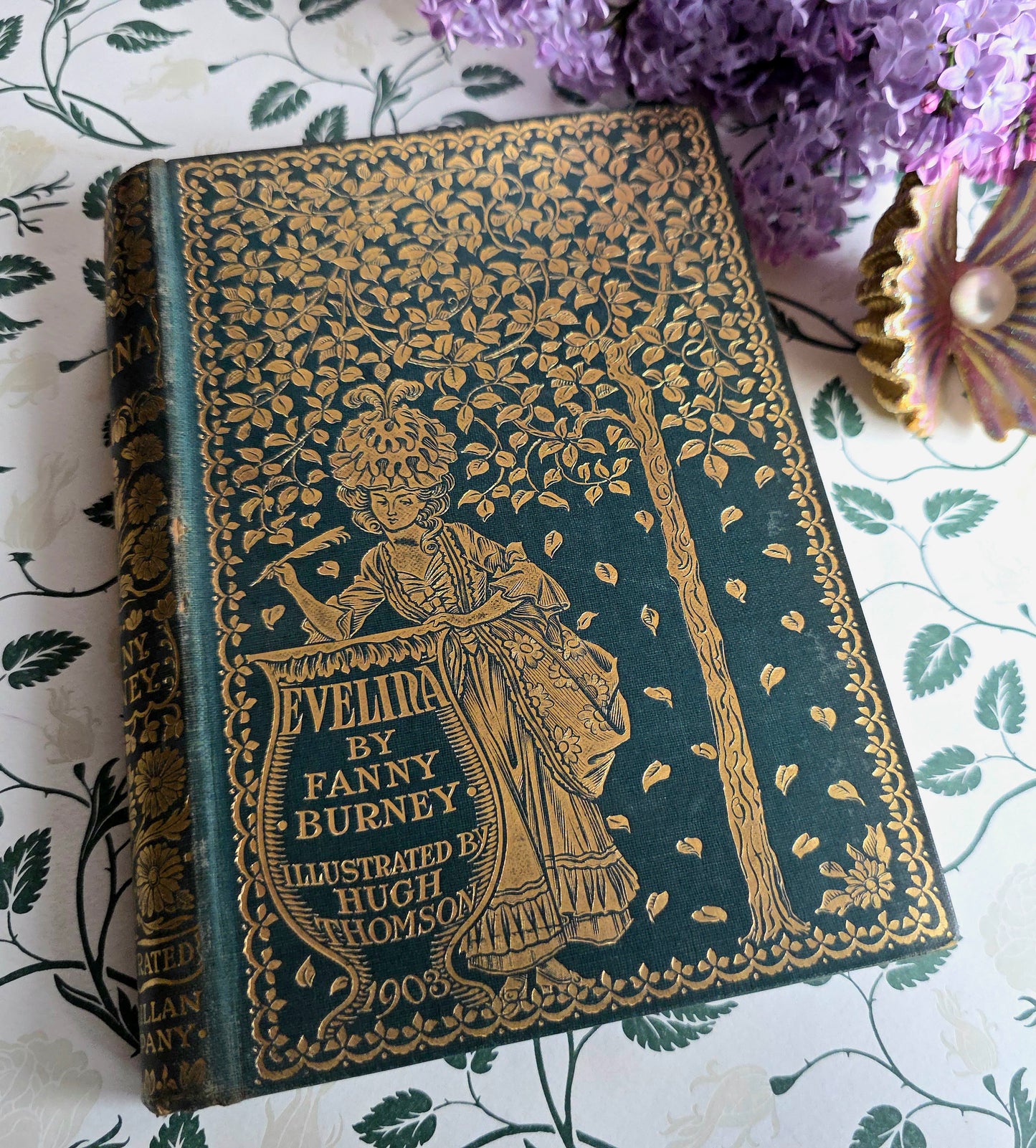 Antique book entitled Evelina by Fanny Burney with gold embossed cover on a floral-patterned surface.