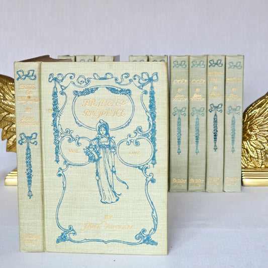 complete set of jane austen novels as a row with one copy placed out front in pale green cloth with blue decoration including a lady holding a basket and gilt titles